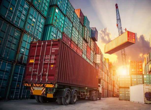 Container truck in a stacked container yard with a crane lifting a box overhead